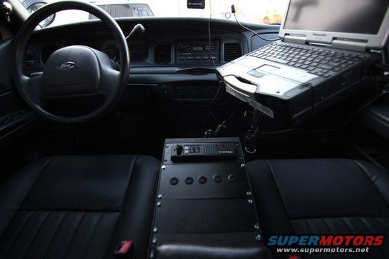 Interior of a 2004 Ford Crown Victoria police car, showing the steering wheel, dashboard, laptop, and radio equipment