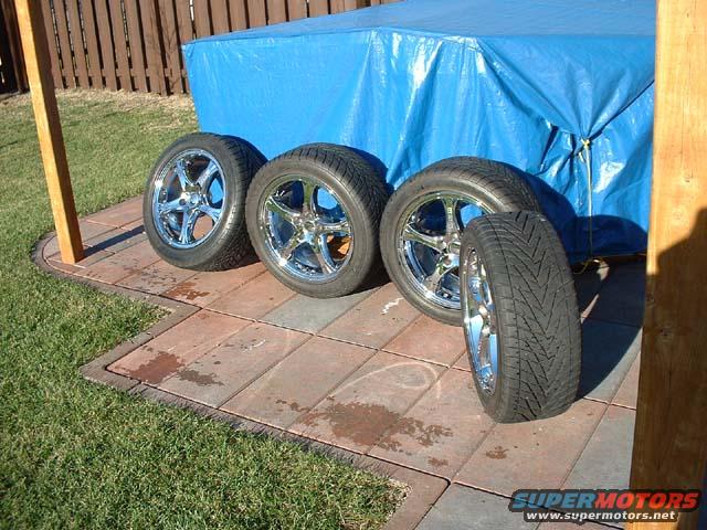 Four chrome wheels with tires are laid out on a patio next to a blue tarp-covered object