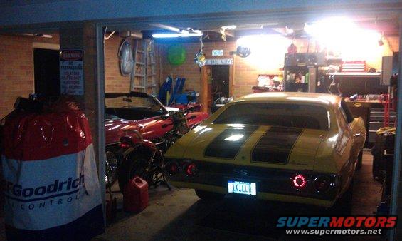 Yellow 1970 Chevrolet Chevelle SS muscle car, rear view, black racing stripes, custom license plate '1KILLAH', parked in a garage