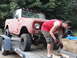 1961 International Scout 80 - A person works on a red off-road vehicle being transported on a trailer, with another vehicle in the background.