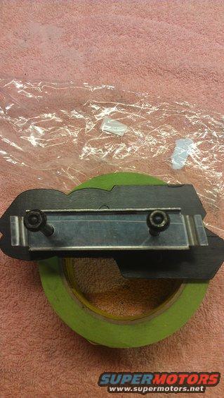 Black automotive emblem with two screws attached to a metal bracket, resting on green tape