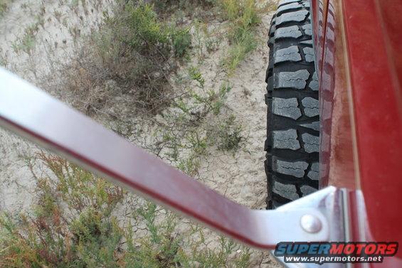 Close-up of an off-road tire and the red side panel of a vehicle, with dry brush and sand in the background. Supermotors.net logo visible