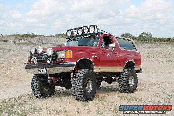 Red lifted Ford Bronco with a light bar on top and extra lights on the front bumper, parked on a sandy, outdoor terrain