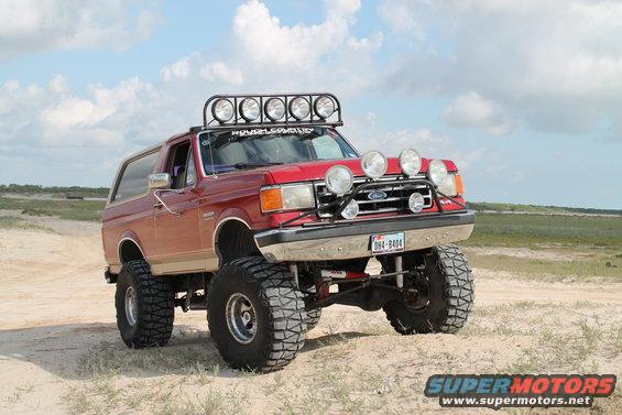 Red 1990 Ford Bronco with off-road lights and lifted suspension on sandy terrain