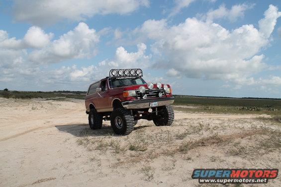 1990 Ford Bronco lifted with off-road lights on sandy terrain under a cloudy sky