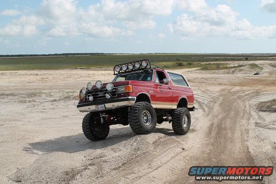 Red 1987 Ford Bronco with lifted suspension and multiple auxiliary lights on a sandy terrain