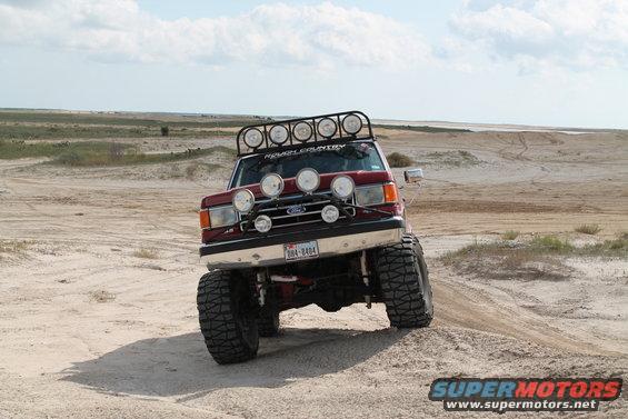 Red 1990 Ford Bronco with roof and bumper lights driving on sand