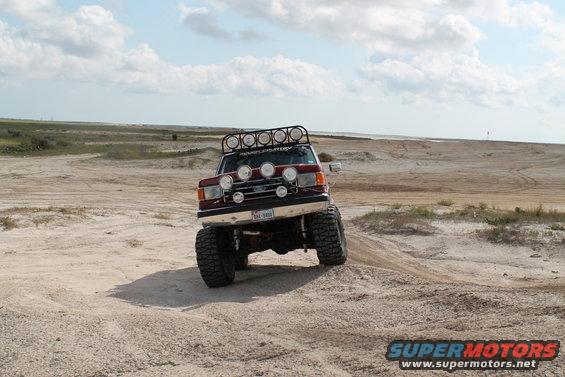 1990 Ford Bronco with off-road lights and roof rack driving on sand dunes