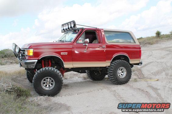 Red 1990 Ford Bronco with tan trim, lifted suspension, and roof-mounted lights on a sandy terrain