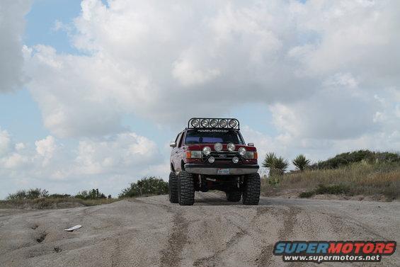 Red Ford Bronco with large tires and roof-mounted lights driving on a dirt road
