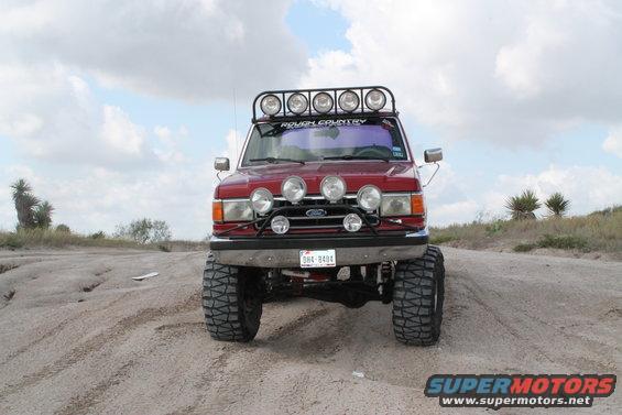 Red Ford Bronco with a light bar on top and extra lights on the front, lifted with large tires, on a dirt road