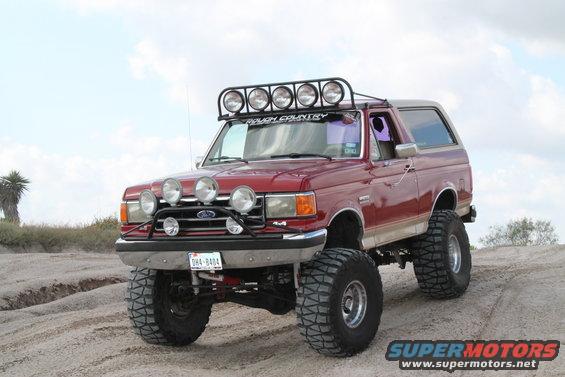 Red 1990 Ford Bronco with off-road lights and lifted suspension on sandy terrain