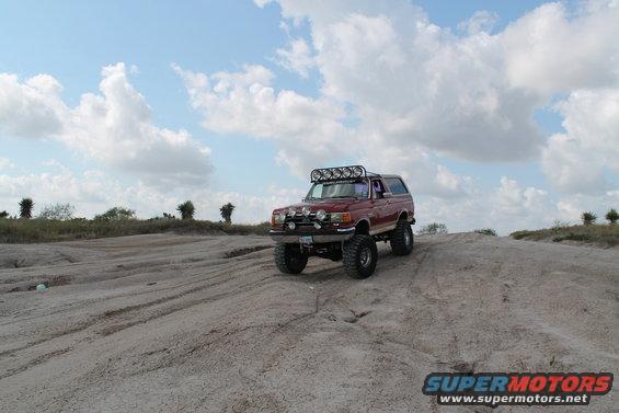 Red 1990 Ford Bronco with off-road lights and large tires driving on a dirt hill