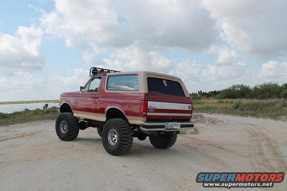 Lifted red Ford Bronco with a tan top parked on a dirt road near water