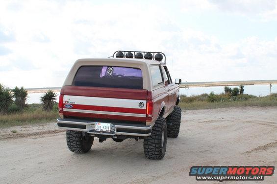 Red and tan 1990 Ford Bronco with lifted suspension and off-road tires, viewed from the rear