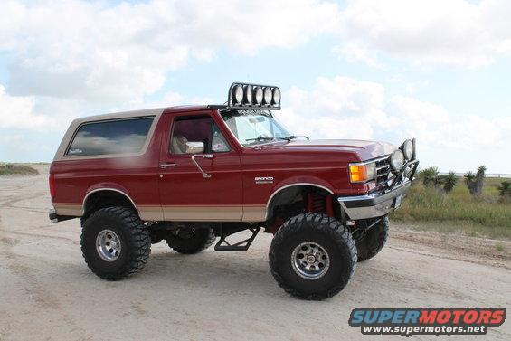 1990 Ford Bronco, red with tan top, lifted suspension, off-road lights on roof and bumper, parked on dirt road near coast