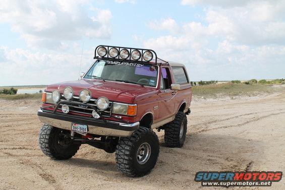 Red 1990 Ford Bronco with roof and bumper lights, lifted on off-road tires