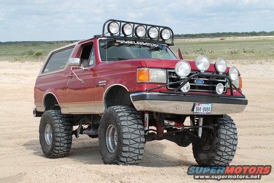 Red 1987 Ford Bronco with lifted suspension, large off-road tires, and a roof-mounted light bar parked on sand