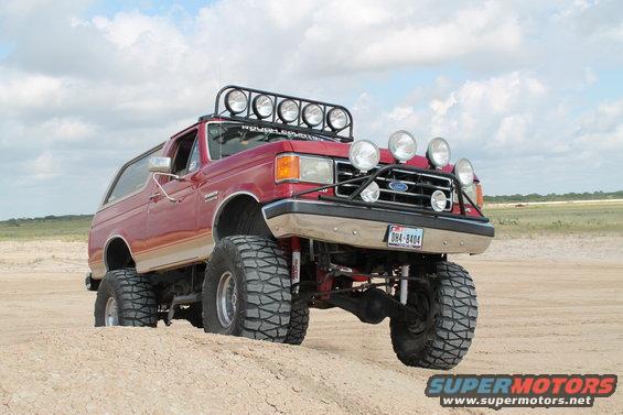 Red 1990 Ford Bronco with large off-road tires and light bar on roof, parked on sand