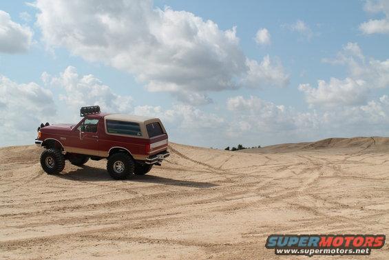 1990 Ford Bronco with lifted suspension and roof rack driving on sand dunes