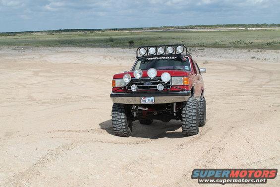 1990 Ford Bronco modified for off-roading, parked on sand with large tires and multiple spotlights