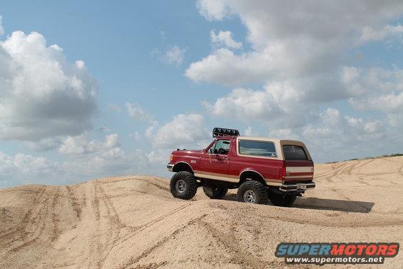 Red 1990 Ford Bronco with lifted suspension driving on sand dunes under a cloudy sky