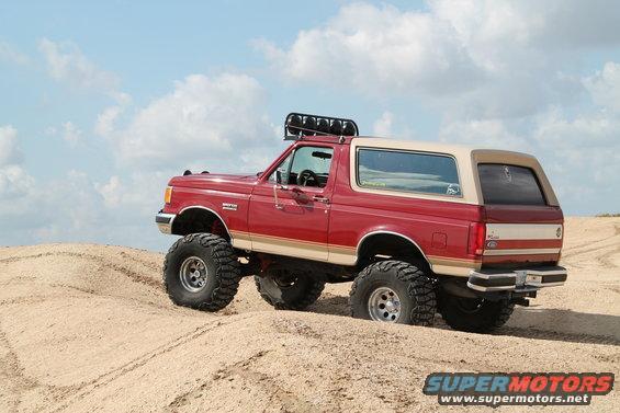 Red 1990 Ford Bronco with tan top and large off-road tires on sand dunes