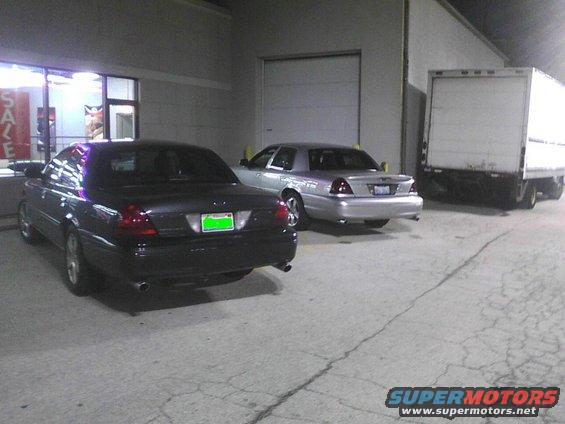 Two Ford Crown Victorias parked outside a building, one dark and one silver, with a box truck to the right