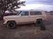 1988 Ford Bronco - A tan Jeep Wagoneer sits parked on a dirt lot under a cloudy sky, with a tree and a tractor visible in the background.