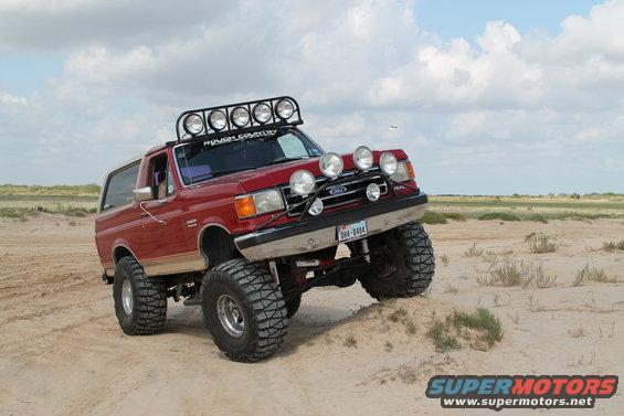 Red 1990 Ford Bronco with off-road lights and roof rack on sandy terrain