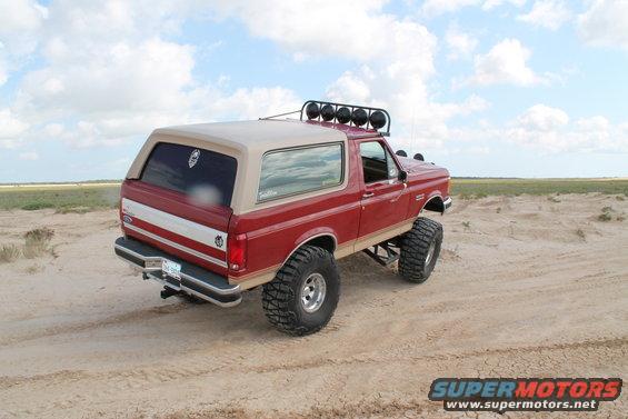 Red 1990 Ford Bronco with tan top, lifted suspension, and off-road tires on sandy terrain