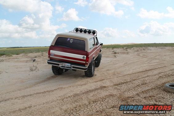 Rear view of a 1990 Ford Bronco with off-road tires and a light bar on a sandy terrain