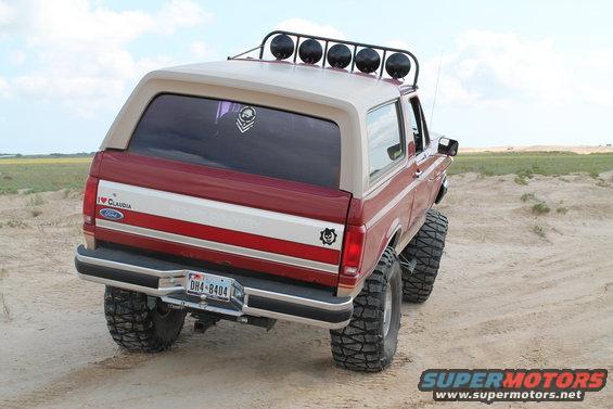 Red Ford Bronco with tan top, lifted on big tires, parked on dirt. Has a light bar on the roof