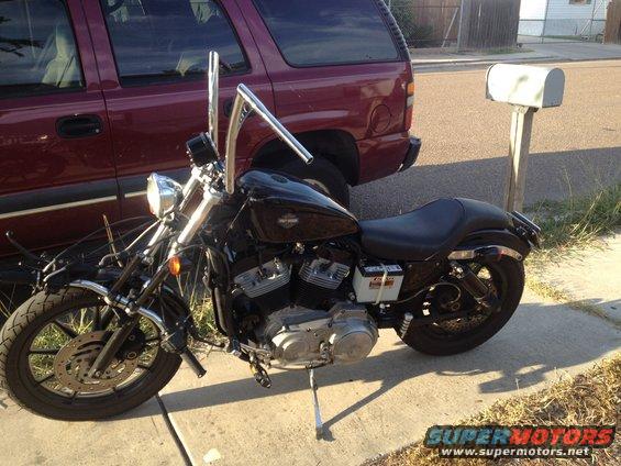 Black Harley-Davidson motorcycle with tall handlebars parked on a sidewalk next to a maroon SUV