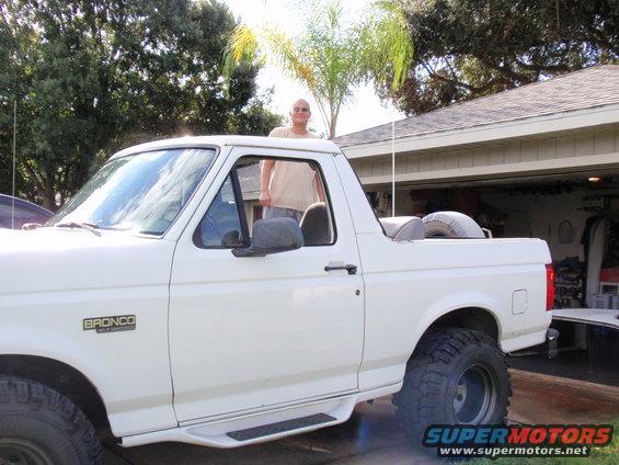 White Ford Bronco with a man in the driver's door, spare tire in the bed, and aftermarket wheels