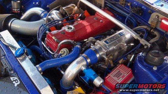Close-up of a blue engine bay with red valve covers, silver intercooler piping, and blue hoses. A red 'Boost Master' box is visible