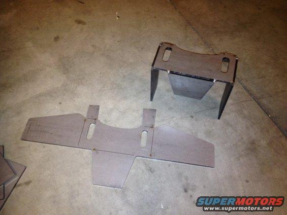 Two unpainted metal automotive parts on a concrete floor. One is flat with cutouts, the other is a bracket