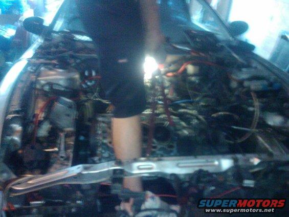 Engine bay with exposed wires and components, a person's legs visible. The car's front end is partially visible