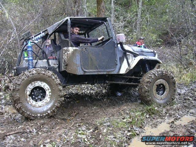 Modified off-road vehicle with large tires driving through mud and trees. Two people are inside