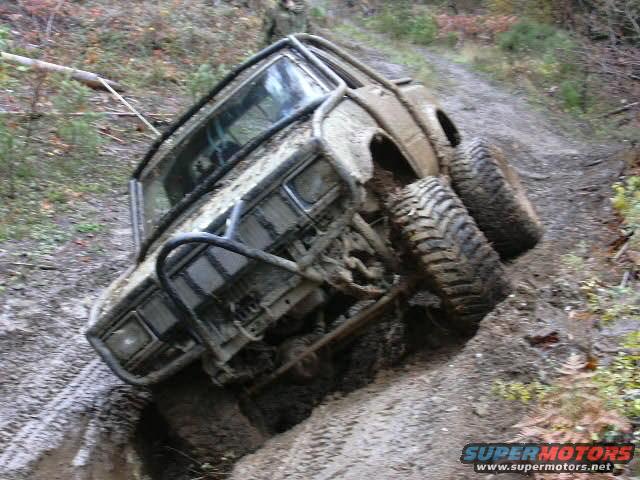 Mud-covered off-road vehicle tilted on a muddy incline, showing large tires and a roll cage