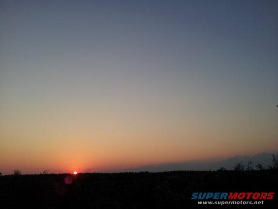 Sunset over a dark landscape with a faint lens flare. SUPERMOTORS.NET logo in the lower right