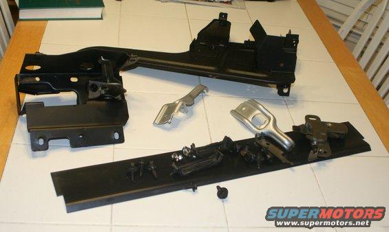 Various black and silver metal vehicle parts, including a large frame-like component and smaller brackets and fasteners, arranged on a tiled surface
