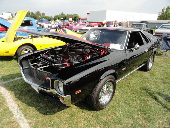 Black AMC Javelin with hood open, showing engine. Yellow car and blue truck visible behind it