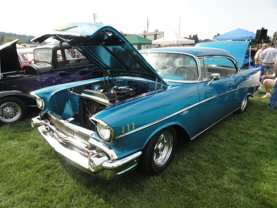 Blue 1957 Chevrolet Bel Air with hood open, parked on grass next to a dark purple car with flame decals