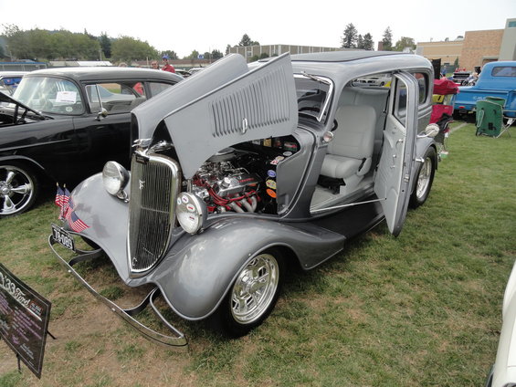 Gray 1933 Ford hot rod with hood and doors open, showing engine and interior. Another car is parked to the left