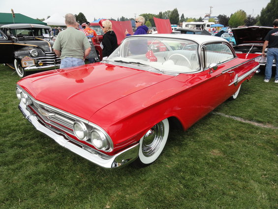 Red and white 1960 Chevrolet Impala with wide whitewall tires and chrome wheels. Other cars and people are visible in the background
