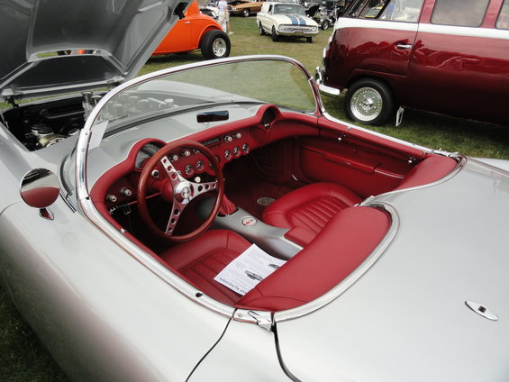 Silver 1950s Corvette convertible interior with red seats and dashboard. Other cars visible in background