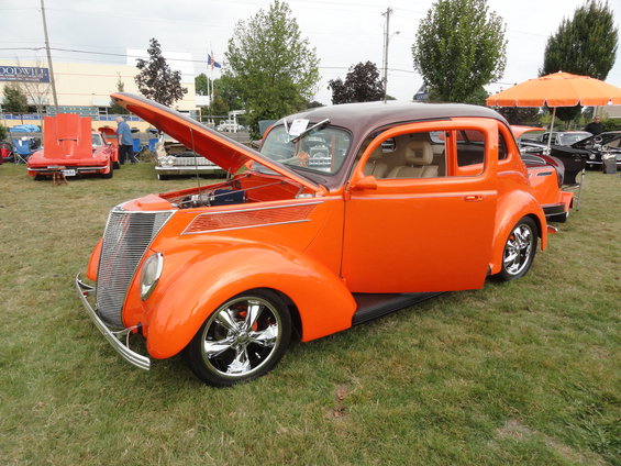 Orange hot rod sedan with hood and door open, parked on grass at a car show. Another orange car and a black car are visible in the background