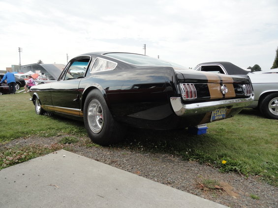 Black 1965 Ford Mustang Fastback with gold stripes, rear view. Another car is parked to its right
