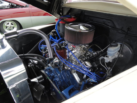 Engine bay with blue spark plug wires, chrome air cleaner, and a shiny radiator. Another car is visible in the background